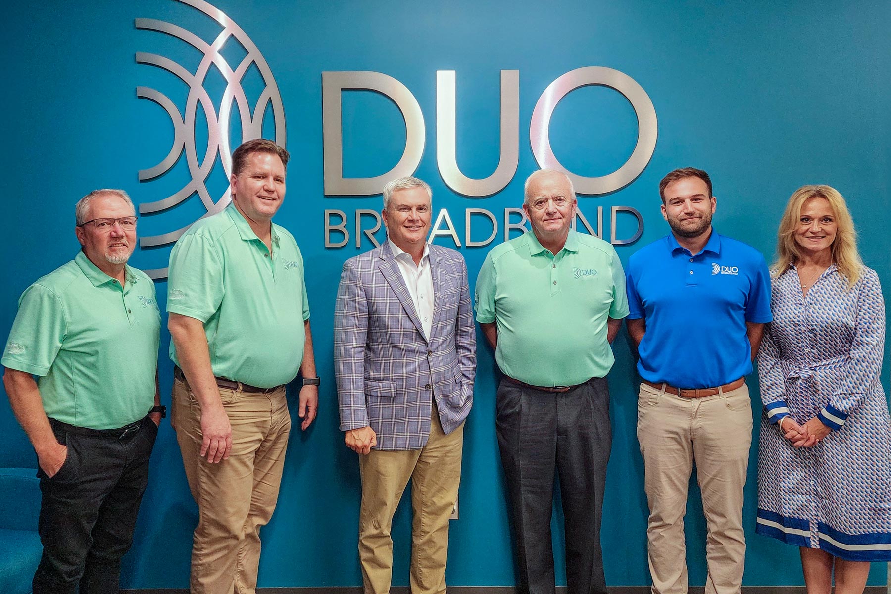 Group of 6 people in front of a wall with DUO Broadband logo